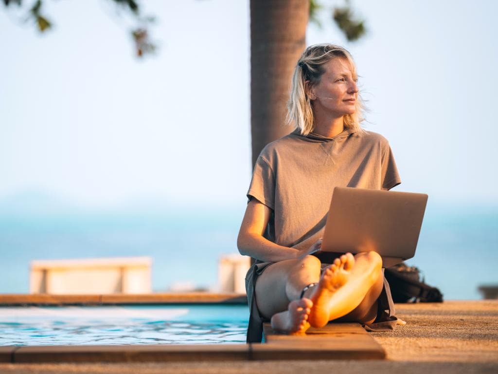 Ženska sedi ob bazenu, dela na prenosniku in uživa v sončnem dnevu na prostem