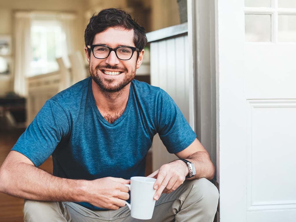 Moški z očali in modro srajco sedi na tleh ter v rokah drži skodelico kave